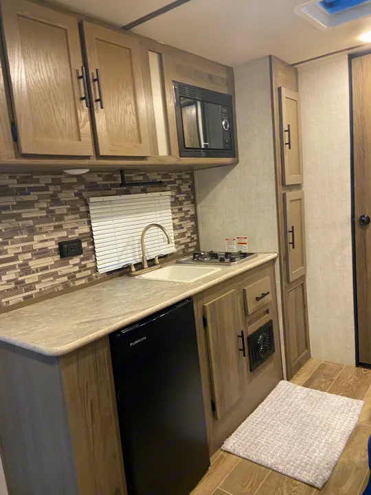 Experience an RV rental's cozy kitchen with a sink, two-burner stovetop, microwave, and Furrion fridge. Features wood cabinets & mosaic backsplash.. Gulf Stream Enlighten 2021