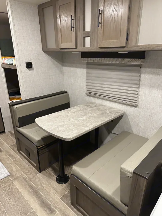 A cozy RV rental dinette with grey benches, a speckled table, overhead cabinets, and glimpse of bunk beds.. Gulf Stream Enlighten 2021
