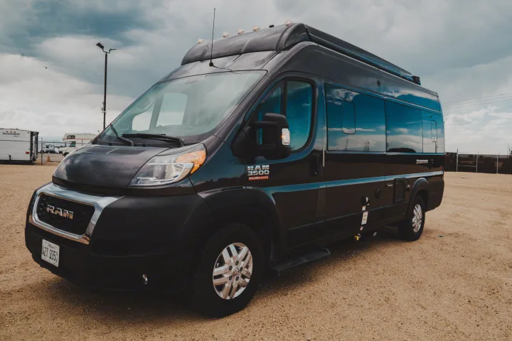 Dark gray Ram Promaster RV rental parked on a gravel lot under cloudy skies.. Thor Motor Coach sequence 2021