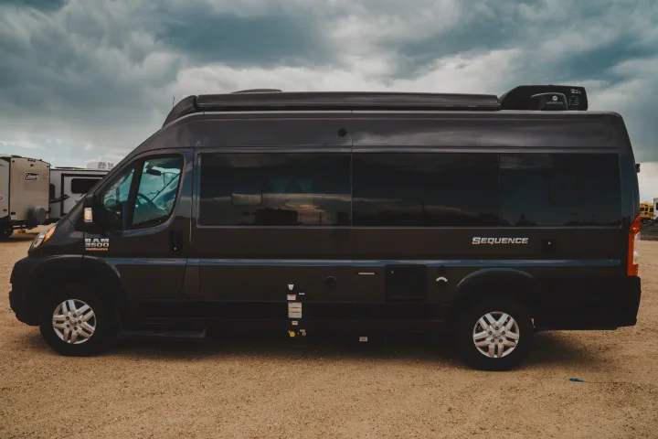 Side view of a dark grey Ram ProMaster "Sequence" Class B RV rental parked on a gravel lot under cloudy skies.. Thor Motor Coach sequence 2021