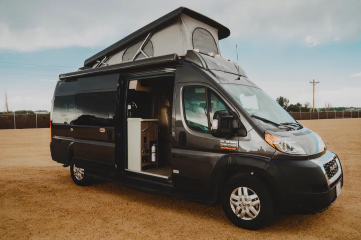 Modern gray Ram ProMaster rv rental camper van with pop-top open, revealing interior kitchenette. Parked on dirt.. Thor Motor Coach sequence 2021