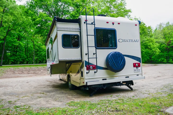 Rear view of a Chateau RV rental, cream-colored with a slide-out, parked on gravel under lush green trees. Great for camping.. Thor Chateau 2017