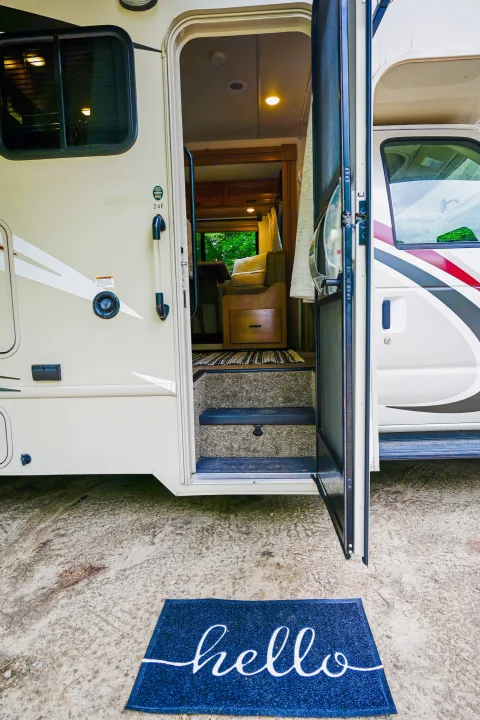 Exterior view of an RV rental with its main door open, showing carpeted steps and a "hello" welcome mat.. Thor Chateau 2017
