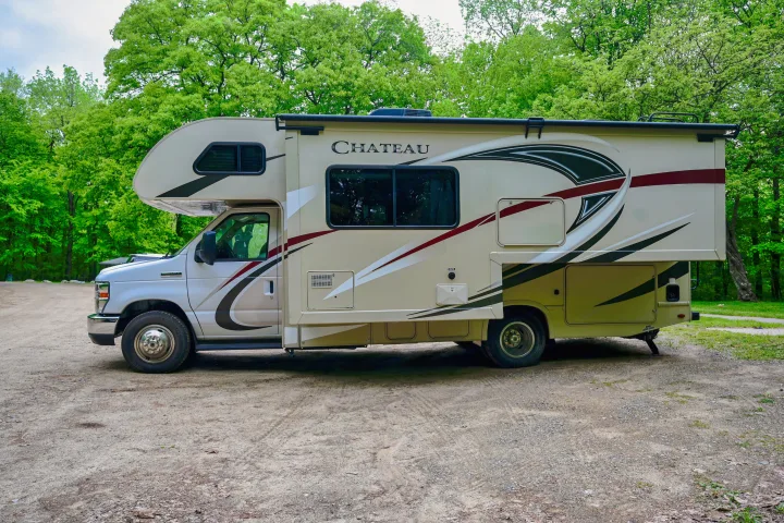 A beige and white Chateau RV rental is parked on a dirt lot, surrounded by lush green trees.. Thor Chateau 2017
