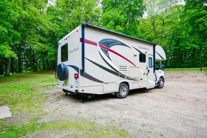 Chateau RV rental parked on a gravel lot, surrounded by vibrant green trees, showcasing its side graphics and back ladder.. Thor Chateau 2017