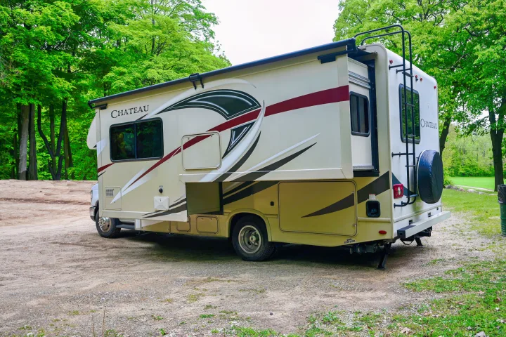 A 'Chateau' RV rental motorhome parked on a dirt road among green trees, featuring an extended slide-out and rear ladder.. Thor Chateau 2017