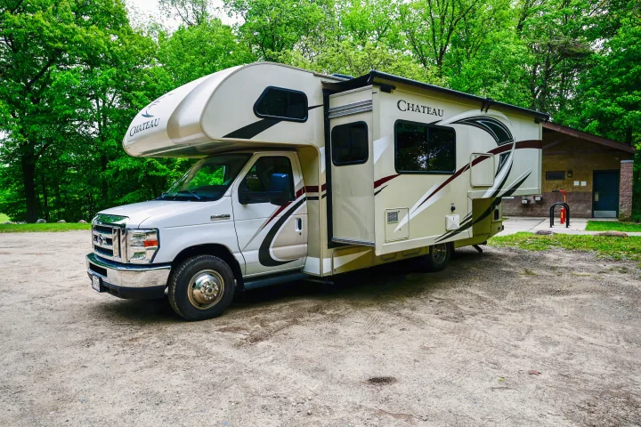 A light-colored Class C Chateau rv rental is parked on a gravel lot, surrounded by vibrant green trees and a park building.. Thor Chateau 2017