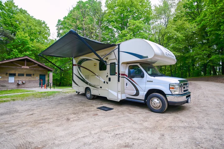 A white Chateau Class C rv rental parked with its awning extended at a wooded campsite next to a utility building.. Thor Chateau 2017