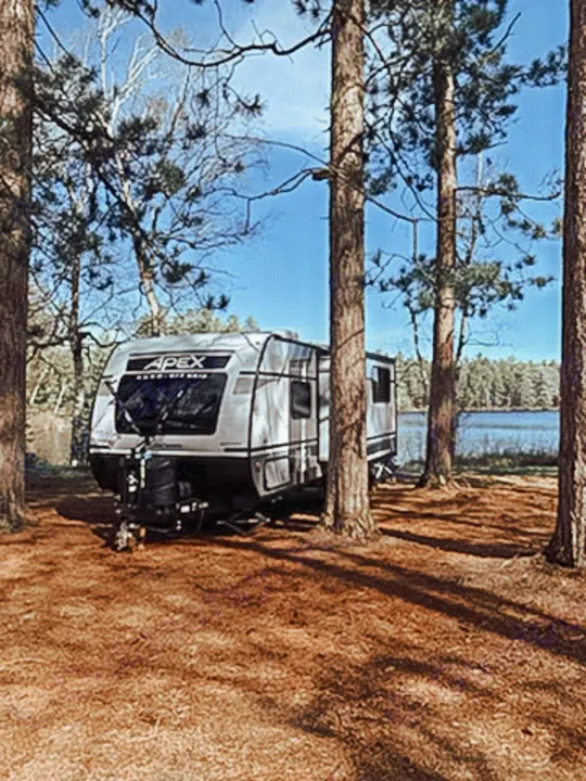 Enjoy nature with an RV rental! This travel trailer is perfectly set up at a serene lakeside campsite among pine trees.. Coachmen Apex Nano 2021