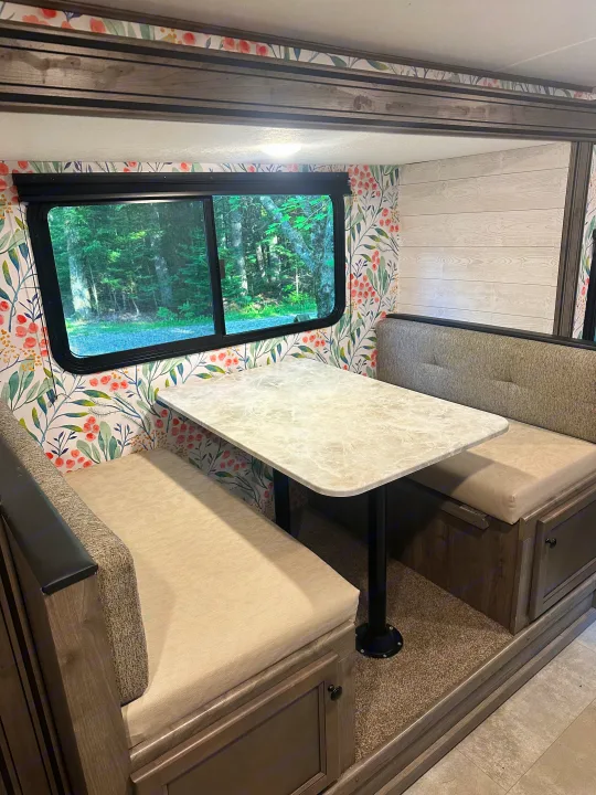 Cozy RV rental dinette with floral wallpaper, comfortable benches, a table, and a scenic forest view outside.. Coachmen Apex Nano 2021