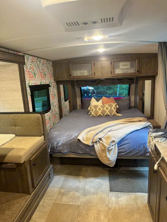 Cozy RV rental interior showcasing a bedroom with a large bed, pillows, and blanket, plus a dinette and floral wallpaper.. Coachmen Apex Nano 2021