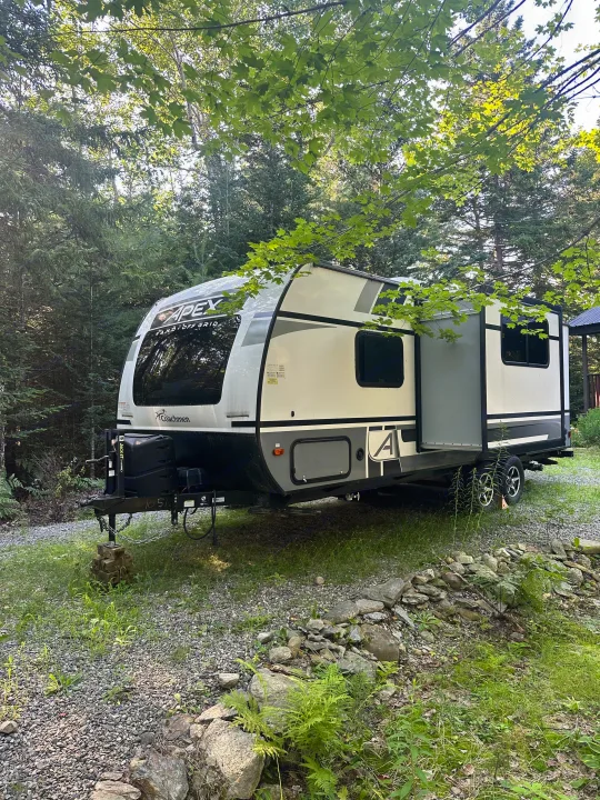 An Apex travel trailer, an rv rental ready for nature. Parked amidst green trees & forest. A white & gray camper with slide-out.. Coachmen Apex Nano 2021