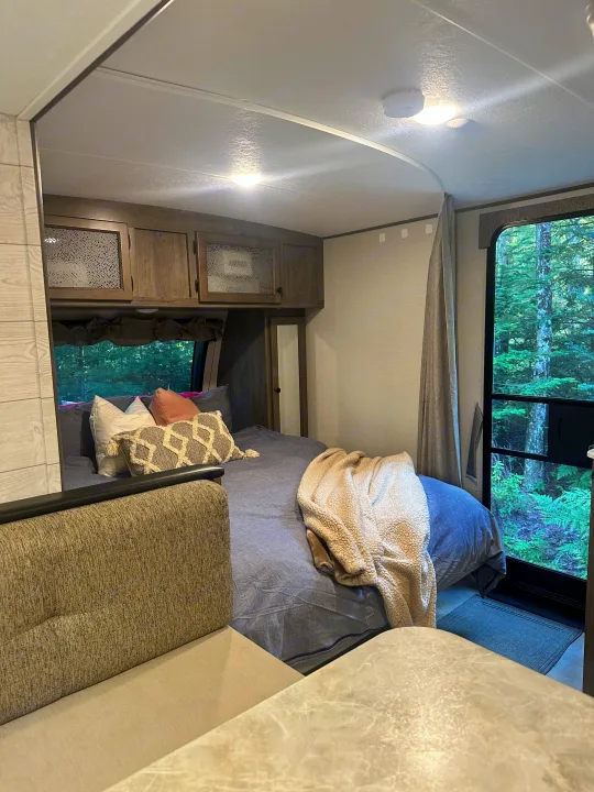 Cozy RV rental interior showcasing a bedroom with a comfy bed, overhead cabinets, and a large window with a lush forest view.. Coachmen Apex Nano 2021