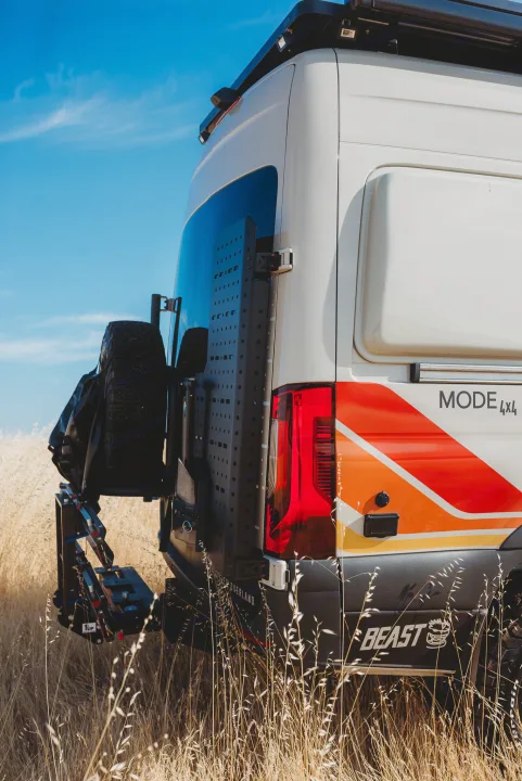 Experience overlanding with this 4x4 rv rental van, featuring custom graphics, rear gear panel, & spare tire in a field.. Storyteller Overland Storyteller Overland Class B 2023
