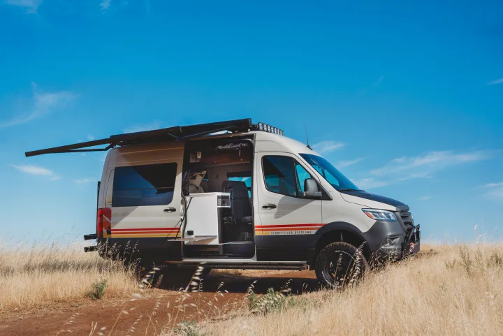 Experience van life! This Storyteller Overland camper van, ideal for an RV rental, is parked off-road with its side door open and awning out.. Storyteller Overland Storyteller Overland Class B 2023