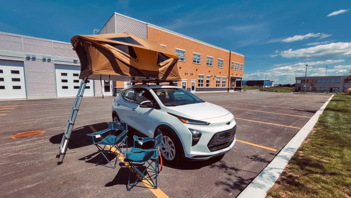 White electric car with roof-top tent & chairs in a parking lot. A compact RV rental alternative for camping.. Chevrolet Other 2023