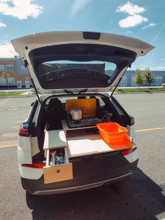 Rear view of a white SUV with an open trunk, revealing a custom RV rental conversion: mobile kitchen, stove, and storage drawers.. Chevrolet Other 2023