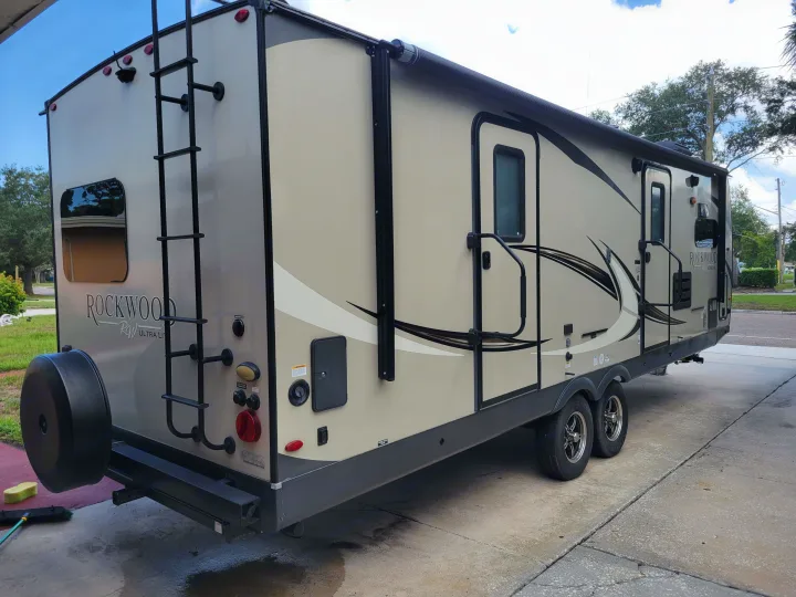 Beige Rockwood RV rental parked on a driveway. Features a rear ladder, spare tire, awning, and multiple access panels.. Forest River Rockwood Ultra Lite 2020