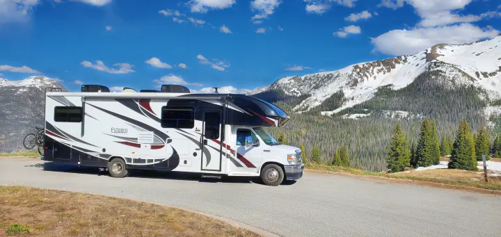An rv rental parked on a mountain road offers stunning views of snow-capped peaks & pine forests under a bright blue sky.. Entegra Coach Esteem 2021