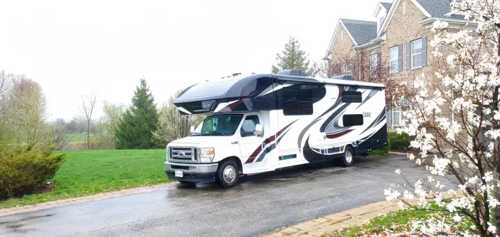 A white Class C Entegra rv rental motorhome with black/maroon trim, parked on a driveway beside a house with a white flowering tree.. Entegra Coach Esteem 2021