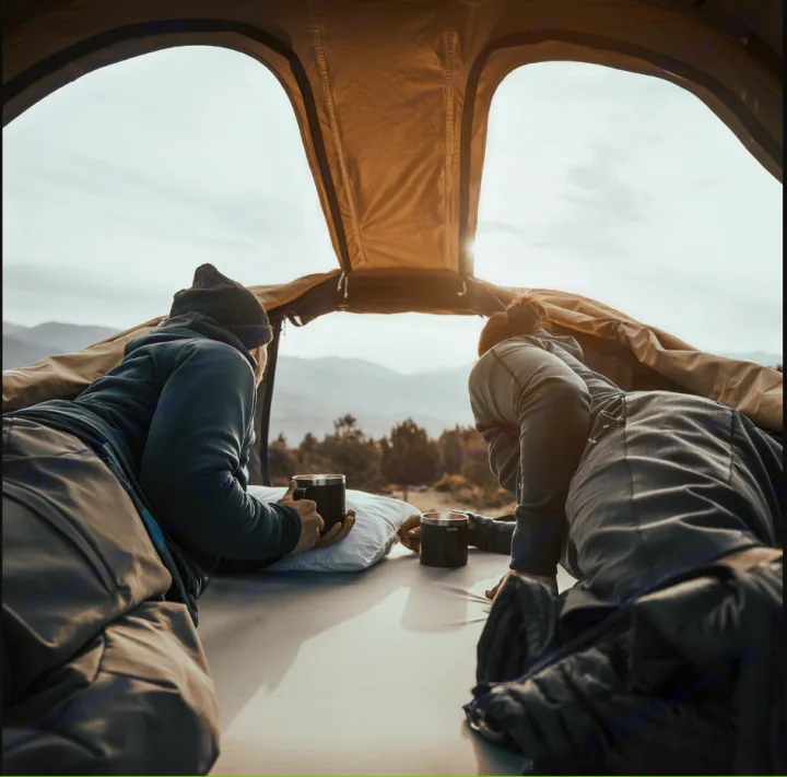 Enjoy peaceful mornings with an RV rental. Two people inside a rooftop tent look out at sunlit mountains, holding coffee mugs.. Chevrolet Other 2023