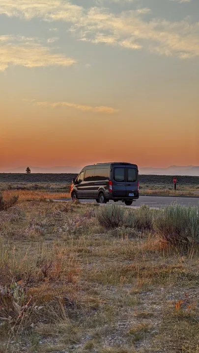 An RV rental van sits on a road at sunset, surrounded by dry fields and distant mountains. A lone tree and stop sign are seen.. Ford Transit 2020