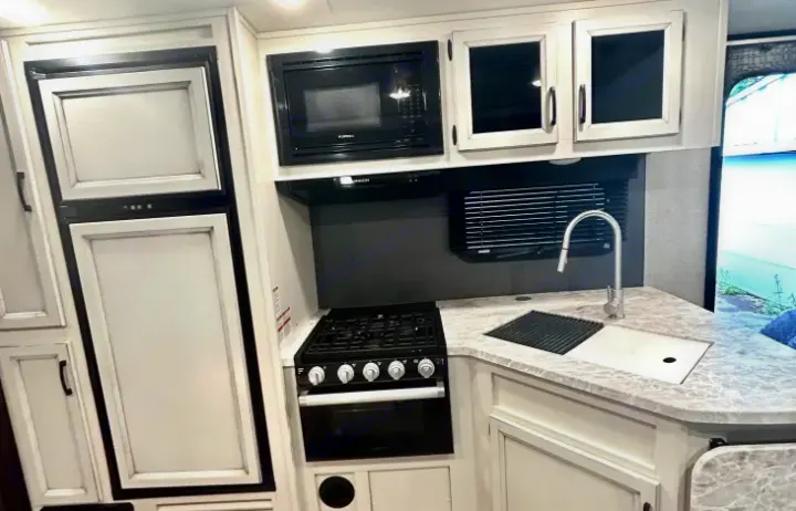 Compact kitchen in an RV rental, featuring a white fridge, black microwave & gas stove, and a sink with drying rack.. Jayco White Hawk 2021