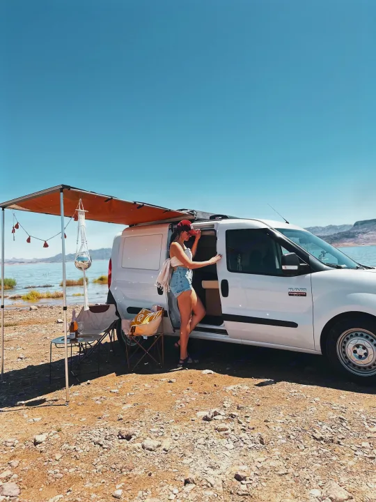 Scenic RV rental adventure: Woman exits white van with awning, camp setup by clear blue lake. Perfect van life!. Dodge Dodge promaster city 2016