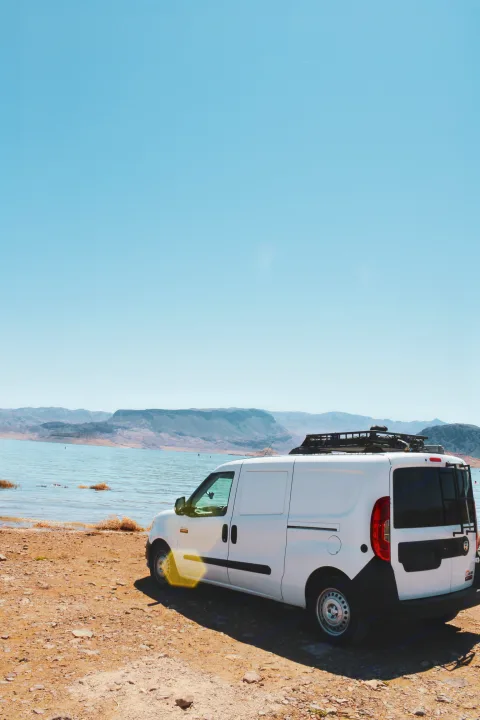 Adventure awaits! White RV rental campervan parked lakeside with mountains under a clear sky.. Dodge Dodge promaster city 2016