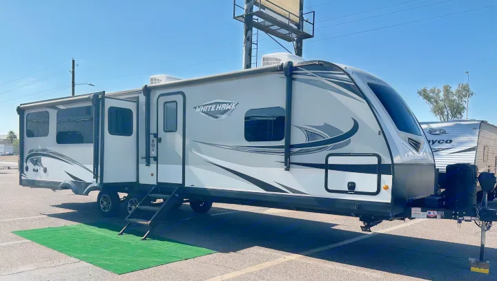 A grey and white White Hawk travel trailer, possibly for RV rental, parked on asphalt with slides extended at a dealership.. Jayco White Hawk 2020