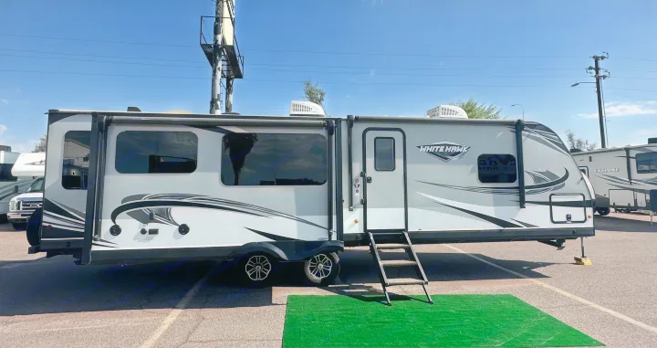 This modern White Hawk RV rental features multiple windows and a front door with steps, parked on asphalt with other RVs nearby.. Jayco White Hawk 2020