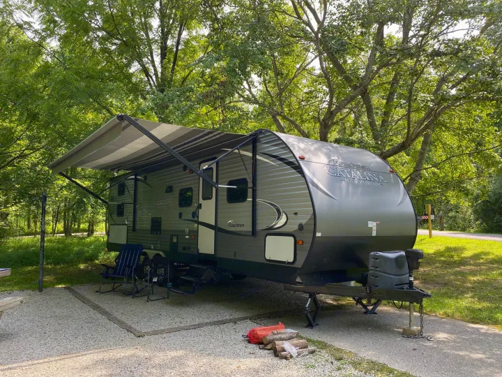 This Coachmen Catalina travel trailer RV rental is parked on a gravel pad at a shaded campground site, with an awning extended.. Coachmen Catalina Legacy 2016