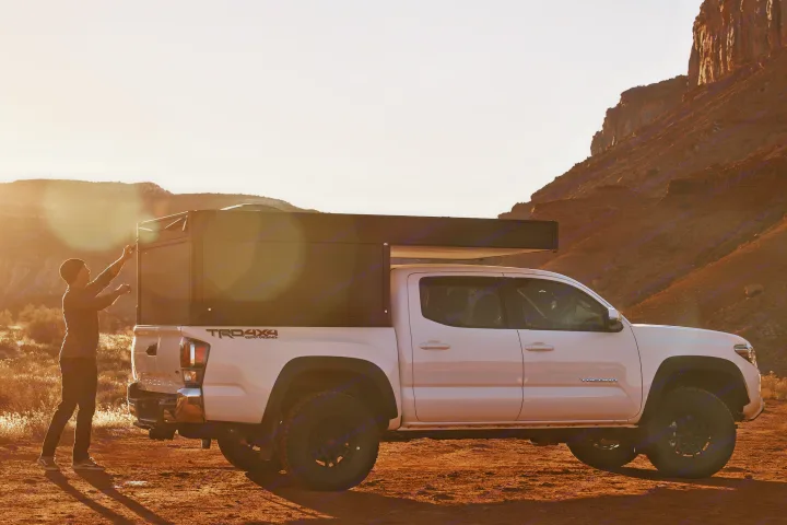 White Toyota Tacoma with pop-up truck camper, being set up by a person in a desert landscape. Perfect for an RV rental adventure!. Toyota Tacoma 2022
