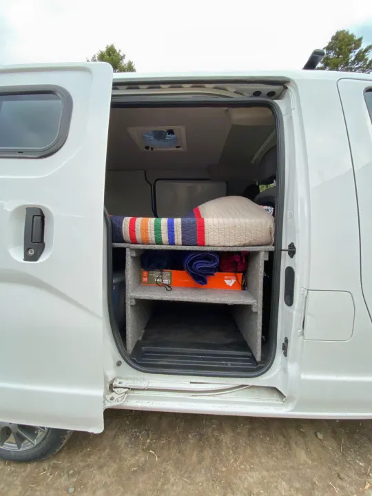 Inside this **RV rental**, a custom van conversion features a raised bed with a striped quilt and smart storage below.. Nissan PRIMASTAR 2014