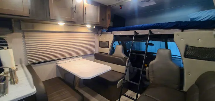 Spacious rv rental interior featuring a dining table, upper bunk bed with ladder, and front cab area. Your road trip awaits!. Coachman Leprechaun 2022