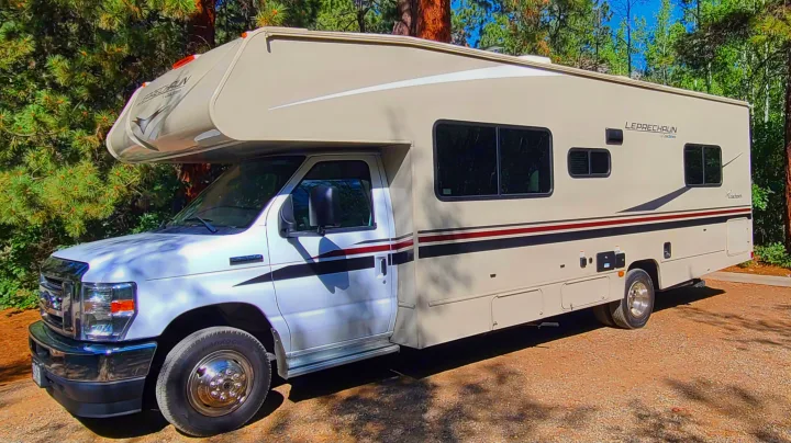Spacious tan Class C **rv rental** parked on gravel, surrounded by green trees under a sunny sky.. Coachman Leprechaun 2022