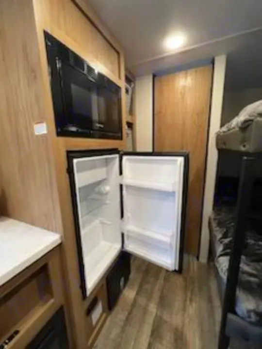 This RV rental interior features a kitchenette with a microwave and an open mini-fridge, plus a glimpse of bunk beds for travel comfort.. Forest River Cherokee 2024