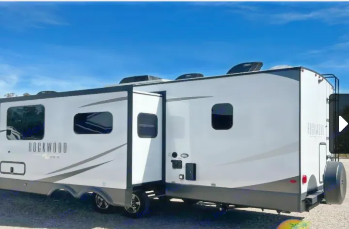 A white and grey Rockwood travel trailer RV rental with an extended slide-out, parked outside under a blue sky.. Forest River Rockwood Ultra Lite 2023