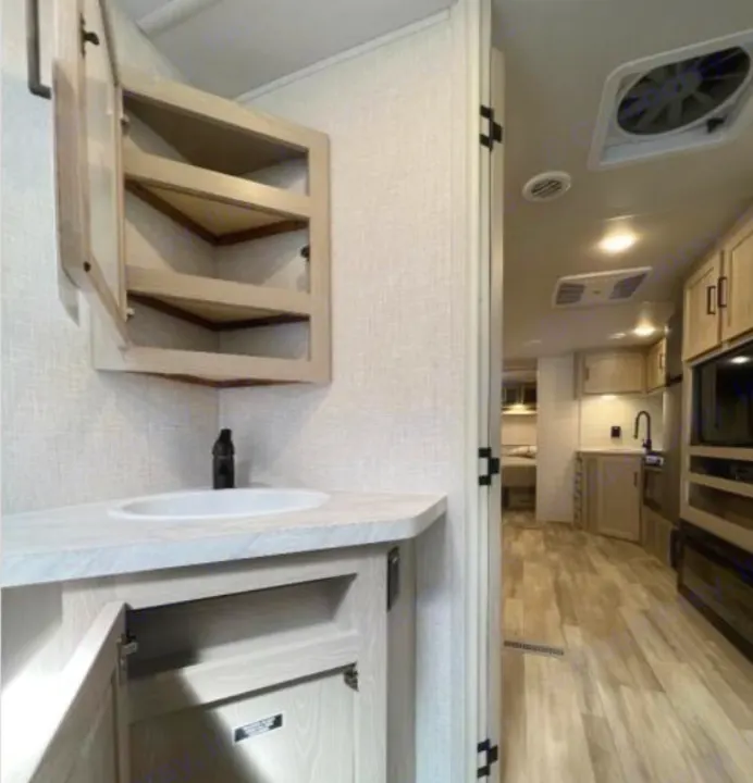 This modern RV rental interior features a light wood bathroom vanity with a sink, a corner medicine cabinet, and a view into the full kitchen area.. Forest River Rockwood Ultra Lite 2023