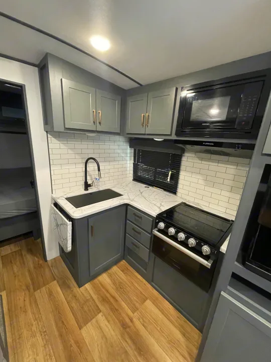 Explore this modern RV rental kitchen, showcasing gray cabinets, white subway tile backsplash, microwave, and range.. Keystone RV Bullet Ultra Lite 2023
