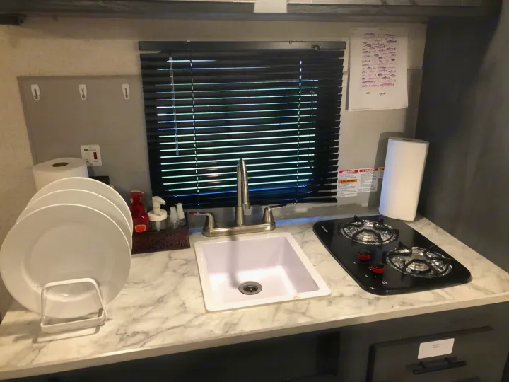 Compact RV rental kitchen with sink, two-burner stove, plates, and counter space. Perfect for camper cooking.. Aspen Trail 17BH 2021