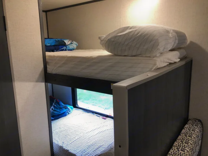 Cozy RV rental bunk beds. The top bunk features white bedding, while the bottom bunk has striped bedding and a window view.. Aspen Trail 17BH 2021