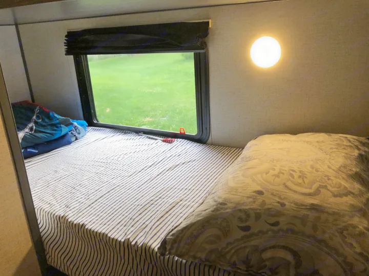 Inside a bright RV rental bedroom: a striped bed, window view of green grass, and a circular wall light. Cozy travel space.. Aspen Trail 17BH 2021