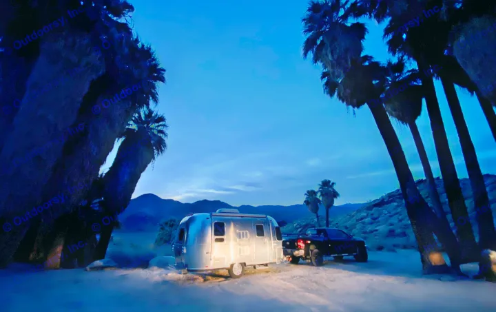An Airstream RV rental & truck set for desert night camping. Interior lights glow under blue twilight, silhouetted palms frame the scene.. Airstream 19 Bambi International 2012