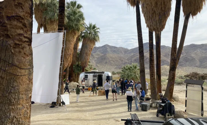 Outdoor shoot featuring an Airstream RV rental in a desert oasis with tall palms & mountains. Crew actively works on set.. Airstream 19 Bambi International 2012