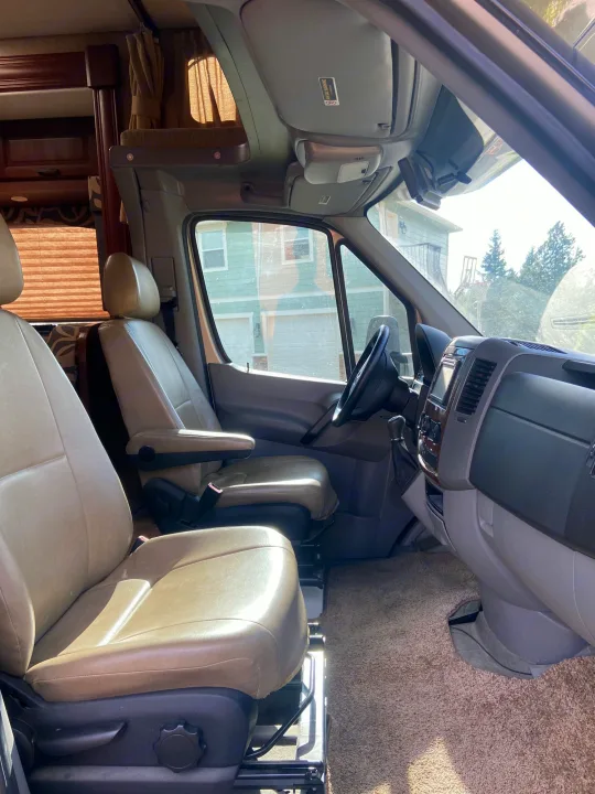 Explore this RV rental's spacious front cabin, featuring comfortable tan seats, a modern dashboard, and an overhead bunk.. Solera Solera Motorhome 2014