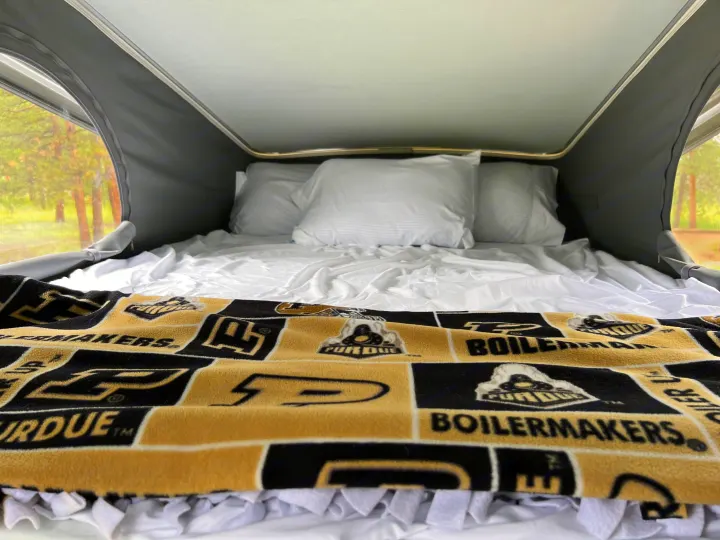 Inside an RV rental pop-top: a comfy bed with white linens and a Purdue Boilermakers blanket, surrounded by nature views.. Winnebago Solis 2023