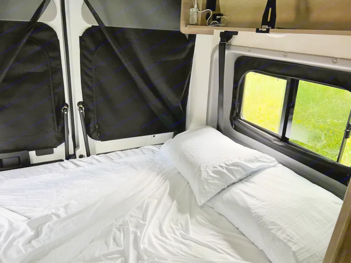 Inside a comfy RV rental: A neat bed with white sheets and pillows next to windows showing a bright green outdoor scene. Ideal for travel rest.. Winnebago Solis 2023