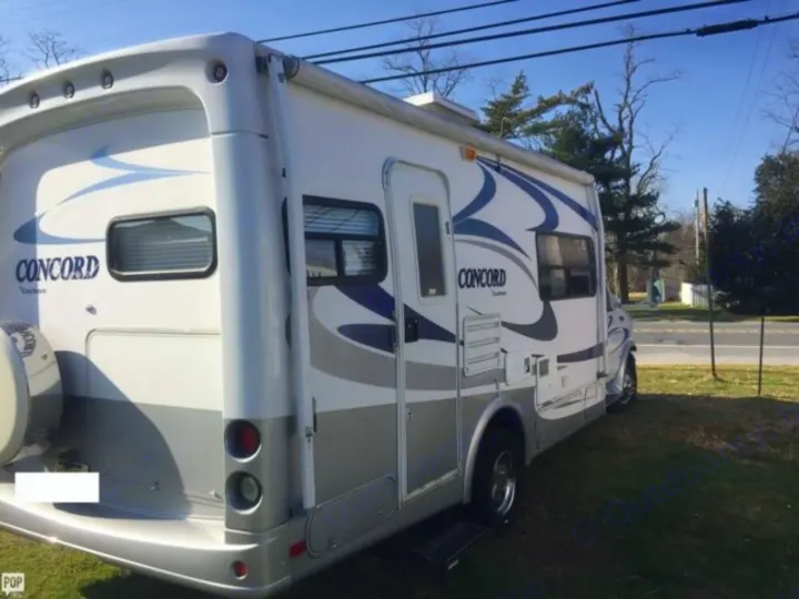 Rear-side view of a white Concord motorhome with blue and gray graphics and an awning, great for an RV rental.. Coachmen Concord 2004