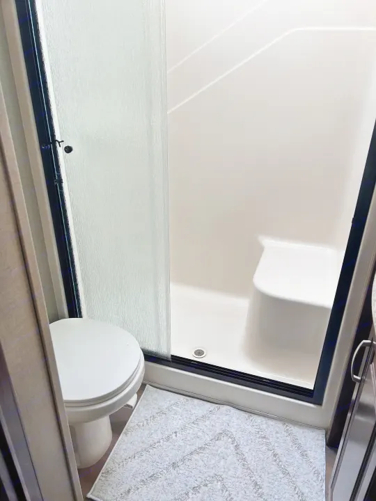 Compact RV rental bathroom featuring a white toilet, frosted glass shower door, built-in shower seat, and a gray rug.. Keystone RV Montana Mountaineer 2015